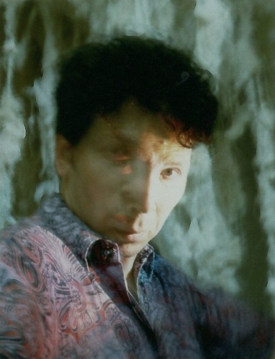Self-portrait of Charly van Rest: half of the face lit, in a patterned shirt, against a pale mottled ground.