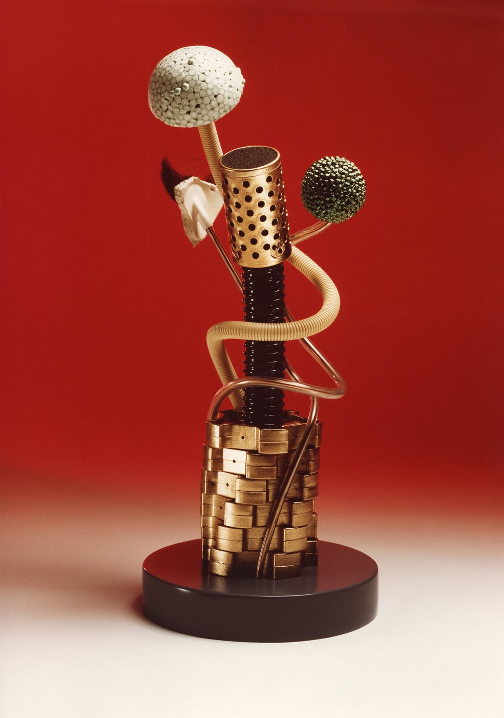 Dogon, 1985 - a totem of gilded brass discs, coiled tube and two seed-head forms rising from a dark round base against a red ground.